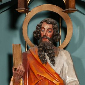 Zdjęcie nr 1: Rzeźba św. Pawła ustawiona na niskim prostopadłościennym cokole o ściętych narożach. Święty ukazany jest frontalnie, w prawej dłoni trzyma księgę, a w lewej miecz skierowany ostrzem do dołu. Twarz o jasnej karnacji, z długim i szerokim nosem, okolona długą i bujną brodą rozdzieloną na dwa pukle, silnie fakturowaną w poziome bruzdy. Włosy ciemnobrązowe, gęste, o bujnych puklach ułożonych po bokach głowy. Ubrany jest w białą suknię z długimi rękawami i kołnierzem oraz jasnobrązowy płaszcz odsłaniający lewe ramię. Wokół głowy ma duży i szeroki, okrągły nimb. Polichromia w odsłoniętych partiach ciała naturalistyczna, detale złocone i srebrzone.
