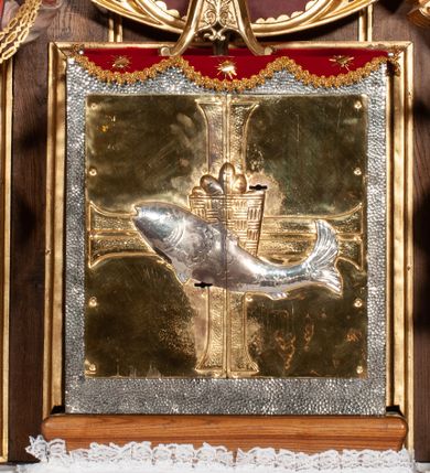 Zdjęcie nr 1: Tabernakulum proste, w formie prostopadłościennej skrzynki, o złotych drzwiczkach z wytłaczaną formą równoramiennego krzyża i wypukłą srebrną rybą oraz koszem z chlebem pośrodku. Po obu stronach, na niewysokich postumentach umieszczone dwa klęczące anioły z wysokimi, uniesionymi do góry skrzydłami, zwróconymi ku środkowi. Twarze o delikatnych rysach, silnie zarumienionych policzkach z 
długimi nosami, głęboko osadzonymi oczami i wysuniętym podbródkiem. Włosy długie, bujne, skręcone w duże pukle po bokach głowy. Anioły ubrane są w białe suknie i czerwone dalmatyki o fioletowych podszewkach. Anioł po lewej stronie trzyma w dłoniach kadzielnicę, a anioł po prawej płomienisty wazon. Polichromie w odsłoniętych partiach ciała naturalistyczne. 



