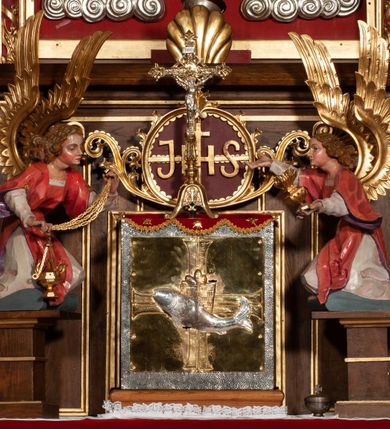 Zdjęcie nr 2: Tabernakulum proste, w formie prostopadłościennej skrzynki, o złotych drzwiczkach z wytłaczaną formą równoramiennego krzyża i wypukłą srebrną rybą oraz koszem z chlebem pośrodku. Po obu stronach, na niewysokich postumentach umieszczone dwa klęczące anioły z wysokimi, uniesionymi do góry skrzydłami, zwróconymi ku środkowi. Twarze o delikatnych rysach, silnie zarumienionych policzkach z 
długimi nosami, głęboko osadzonymi oczami i wysuniętym podbródkiem. Włosy długie, bujne, skręcone w duże pukle po bokach głowy. Anioły ubrane są w białe suknie i czerwone dalmatyki o fioletowych podszewkach. Anioł po lewej stronie trzyma w dłoniach kadzielnicę, a anioł po prawej płomienisty wazon. Polichromie w odsłoniętych partiach ciała naturalistyczne. 


