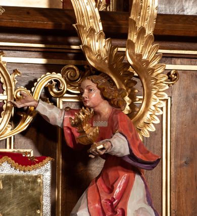 Zdjęcie nr 5: Tabernakulum proste, w formie prostopadłościennej skrzynki, o złotych drzwiczkach z wytłaczaną formą równoramiennego krzyża i wypukłą srebrną rybą oraz koszem z chlebem pośrodku. Po obu stronach, na niewysokich postumentach umieszczone dwa klęczące anioły z wysokimi, uniesionymi do góry skrzydłami, zwróconymi ku środkowi. Twarze o delikatnych rysach, silnie zarumienionych policzkach z 
długimi nosami, głęboko osadzonymi oczami i wysuniętym podbródkiem. Włosy długie, bujne, skręcone w duże pukle po bokach głowy. Anioły ubrane są w białe suknie i czerwone dalmatyki o fioletowych podszewkach. Anioł po lewej stronie trzyma w dłoniach kadzielnicę, a anioł po prawej płomienisty wazon. Polichromie w odsłoniętych partiach ciała naturalistyczne. 


