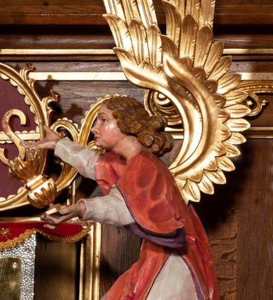 Zdjęcie nr 6: Tabernakulum proste, w formie prostopadłościennej skrzynki, o złotych drzwiczkach z wytłaczaną formą równoramiennego krzyża i wypukłą srebrną rybą oraz koszem z chlebem pośrodku. Po obu stronach, na niewysokich postumentach umieszczone dwa klęczące anioły z wysokimi, uniesionymi do góry skrzydłami, zwróconymi ku środkowi. Twarze o delikatnych rysach, silnie zarumienionych policzkach z 
długimi nosami, głęboko osadzonymi oczami i wysuniętym podbródkiem. Włosy długie, bujne, skręcone w duże pukle po bokach głowy. Anioły ubrane są w białe suknie i czerwone dalmatyki o fioletowych podszewkach. Anioł po lewej stronie trzyma w dłoniach kadzielnicę, a anioł po prawej płomienisty wazon. Polichromie w odsłoniętych partiach ciała naturalistyczne. 


