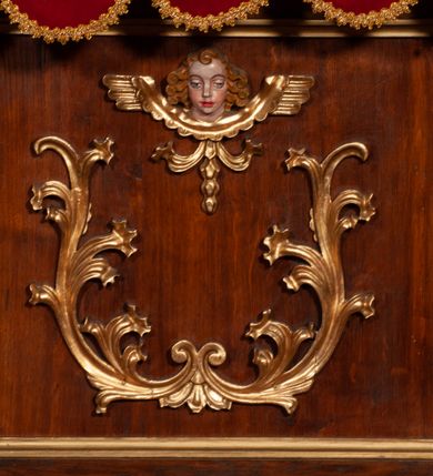 Zdjęcie nr 23: Tabernakulum proste, w formie prostopadłościennej skrzynki, o złotych drzwiczkach z wytłaczaną formą równoramiennego krzyża i wypukłą srebrną rybą oraz koszem z chlebem pośrodku. Po obu stronach, na niewysokich postumentach umieszczone dwa klęczące anioły z wysokimi, uniesionymi do góry skrzydłami, zwróconymi ku środkowi. Twarze o delikatnych rysach, silnie zarumienionych policzkach z 
długimi nosami, głęboko osadzonymi oczami i wysuniętym podbródkiem. Włosy długie, bujne, skręcone w duże pukle po bokach głowy. Anioły ubrane są w białe suknie i czerwone dalmatyki o fioletowych podszewkach. Anioł po lewej stronie trzyma w dłoniach kadzielnicę, a anioł po prawej płomienisty wazon. Polichromie w odsłoniętych partiach ciała naturalistyczne. 


