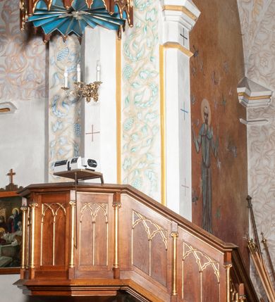 Zdjęcie nr 2: Ambona stojąca, przyścienna, usytuowana przy ścianie północnej, pomiędzy pierwszym a drugim przęsłem nawy. Ustawiona na wysokim, sześciobocznym dwukondygnacyjnym filarze na wysokim cokole. Filar artykułowany smukłymi, złoconymi kolumienkami o jońskich kapitelach. W polach pomiędzy nimi płyciny zamknięte ostrołukowo. Kosz sześcioboczny o stożkowatej podstawie zamkniętej profilowanym gzymsem. Ścianki rozczłonkowane za pomocą analogicznych jak wcześniej kolumienek; w polach prostokątne płyciny, w których przecinające się, złocone arkadki. Całość zamyka profilowany parapet. Baldachim na planie analogicznym do kosza, węższy od niego, od dołu ujęty krenelażem, od góry rzeźbionym, ażurowym ornamentem ze spiętych arkadek, oddzielonych niewielkimi pinaklami. Całość zamknięta architektoniczną strukturą z pinaklem i łacińskim krzyżykiem u szczytu. U podniebienia na niebieskim tle rzeźbiona gołębica Ducha Świętego otoczona promieniami. Na ambonę prowadzą od wschodu, drewniane dwudzielne schody z balustradą dekorowaną analogicznie jak kosz.