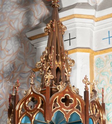 Zdjęcie nr 3: Ambona stojąca, przyścienna, usytuowana przy ścianie północnej, pomiędzy pierwszym a drugim przęsłem nawy. Ustawiona na wysokim, sześciobocznym dwukondygnacyjnym filarze na wysokim cokole. Filar artykułowany smukłymi, złoconymi kolumienkami o jońskich kapitelach. W polach pomiędzy nimi płyciny zamknięte ostrołukowo. Kosz sześcioboczny o stożkowatej podstawie zamkniętej profilowanym gzymsem. Ścianki rozczłonkowane za pomocą analogicznych jak wcześniej kolumienek; w polach prostokątne płyciny, w których przecinające się, złocone arkadki. Całość zamyka profilowany parapet. Baldachim na planie analogicznym do kosza, węższy od niego, od dołu ujęty krenelażem, od góry rzeźbionym, ażurowym ornamentem ze spiętych arkadek, oddzielonych niewielkimi pinaklami. Całość zamknięta architektoniczną strukturą z pinaklem i łacińskim krzyżykiem u szczytu. U podniebienia na niebieskim tle rzeźbiona gołębica Ducha Świętego otoczona promieniami. Na ambonę prowadzą od wschodu, drewniane dwudzielne schody z balustradą dekorowaną analogicznie jak kosz.