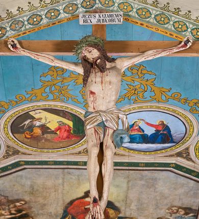 Zdjęcie nr 2: Figura Chrystusa Ukrzyżowanego w lekkim zwisie, o szeroko rozłożonych ramionach tworzących kąt rozwarty. Głowa przechylona na prawe ramię. Twarz pociągła, szczupła, okolona zarostem. Oczy zamknięte, prosty i długi nos, lekko rozchylone usta. Włosy brązowe w formie grubych, spiralnie skręconych pukli, opadających na prawe ramię. Broda krótka, kędzierzawa. Korona cierniowa w kształcie czepca w kolorze zielonym. Sylwetka ciała wychudzona z nienaturalnie długimi, szczupłymi ramionami. Na ciele widoczne ślady męki – krwawiąca rana w boku oraz dłoniach i stopach. Perizonium srebrno-niebieskie, ściśle opinające biodra, przewiązane z przodu, opadające ostro łamaną fałdą wzdłuż lewego boku postaci. Polichromia ciała naturalistyczna. Krzyż gładki, prosty w kolorze jasnobrązowym. U szczytu pionowej belki krzyża titulus z napisem „JEZUS NAZARENUS / REX JUDAEORUM”.