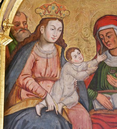 Zdjęcie nr 2: Obraz w kształcie stojącego prostokąta zamkniętego łukiem półkolistym. W centrum kompozycji ukazane frontalnie, siedzące obok siebie Maria trzymająca Dzieciątko na kolanach (z lewej) oraz św. Anna (z prawej). Maria przedstawiona jako młoda kobieta o owalnej twarzy oraz długich, falowanych włosach w kolorze jasnobrązowym, opadających na ramiona. Jej głowa ujęta w trzech czwartych zwrócona jest w kierunku św. Anny i nieznacznie pochylona w dół. Oczy ciemne, wzrok skierowany do dołu, w kierunku Dzieciątka. Nos wydatny, prosty, usta niewielkie, jasnoróżowe. Karnacja postaci jasna z delikatnie zaznaczonymi różem policzkami. Maria ubrana jest w jasnoróżową suknię z długimi rękawami przewiązaną w pasie jasnobrązową szarfą oraz obszerny, niebieski płaszcz z żółtą podszewką. Wokół dekoltu biała chusta. Spod szat widoczna jest stopa obuta w czerwony sandał. Na głowie postaci znajduje się wysadzana kamieniami, złota korona. Wokół głowy nimb zaznaczony w złoconym tle. Dzieciątko zostało ukazane w dynamicznej pozie, klęczy na kolanach Marii, z rękami wyciągniętymi w kierunku św. Anny. Jego okrągła twarz ma jasną karnację z wyraźnie zaznaczonym rumieńcem. Oczy są ciemne, nos prosty, a usta małe. Włosy Dzieciątka są krótkie, jasne i kręcone. Postać ubrana jest w tunikę z długimi rękawami wykonaną z przejrzystej tkaniny. W górnej części kompozycji, w partii tła, wysoko ponad głową Dzieciątka wyryta korona. Święta Anna została przedstawiona jako dojrzała kobieta o wyrazistych rysach twarzy z dużymi, ciemnymi oczami oraz wydatnym nosem. Głowę pochyla w dół, a wzrok zwraca w kierunku Dzieciątka. W swojej lewej dłoni trzyma niewielki kwiat (różę?), prawą podtrzymuje otwartą księgę. Święta ubrana jest w ciemnozieloną suknię z długimi rękawami, wokół szyi oraz na głowie nosi białą chustę, na którą narzucony jest czerwony płaszcz o jasnobrązowej podszewce. Spod szat widoczna jest stopa obuta w czarny trzewik o lekko spiczastym nosku. Wokół głowy świętej zaznaczony w złoconym tle nimb. Na drugim planie, w nieco pomniejszonej skali ukazani zostali św. Józef (z lewej strony obrazu) oraz św. Joachim (z prawej). Święty Józef został przedstawiony jako starszy siwy mężczyzna z włosami przerzedzonymi w okolicach czoła oraz z długą, siwą brodą i wąsami. Oczy duże, ciemne, nos krótki, na czole postaci delikatnie zaznaczone zmarszczki. Postać ubrana w zielone i brązowe szaty. Świętego Joachima ukazano jako starszego mężczyznę o podłużnej twarzy, dużych, ciemnych oczach, wydatnym, prostym nosie z długą, siwą brodą i wąsami. Ubrany jest w granatowe szaty oraz czerwony kapelusz. Tło obrazu złocone, dekorowane rytym ornamentem roślinnym o symetrycznym przebiegu. W dolnej części obrazu, na pierwszym planie przedstawione białe, czerwone oraz niebieskie kwiaty o zielonych łodyżkach ukazane na brązowym tle.