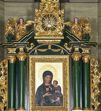 Zdjęcie nr 8: Obraz w kształcie stojącego prostokąta ujęty profilowaną ramą zdobioną akantem. W centrum kompozycji ukazana Matka Boska z Dzieciątkiem w typie Hodegetrii. Maria przedstawiona frontalnie, w popiersiu z głową nieznacznie pochyloną w lewo, w kierunku Dzieciątka, które trzyma na lewym ramieniu. Prawą ręką wskazuje na Jezusa. Twarz Matki Boskiej szczupła, z długim, prostym nosem i niewielkimi ustami. Na policzkach zaznaczony delikatny rumieniec. Wokół głowy złocony nimb z umieszczoną w otoku gotycką inskrypcją: „Regina • celi • letare • alle • quia • quem • meruisti • portare • alle • resurrexit • sicu”. Maria ubrana jest w ciemnogranatową szatę spodnią z długimi rękawami, wykończoną złoconą lamówką. Przy dekolcie gotycki, minuskułowy napis: „ave • maria”. Na ramiona i głowę ma narzucony ciemnogranatowy płaszcz z purpurową podszewką, spięty pod szyją ozdobną, kameryzowaną broszą. Spod płaszcza widoczna przejrzysta chusta oraz jasne, falowane włosy. Dzieciątko Jezus zwrócone w stronę Marii, prawą rękę wyciąga w geście błogosławieństwa, w lewej zaś trzyma zamkniętą księgę w zielonej oprawie. Przedstawione jako brązowooki chłopiec o krótkich, jasnych i kręconych włosach. Dzieciątko ubrane jest w ciemnogranatową tunikę z długimi rękawami zdobioną motywem kwiatowym, wykończoną złotą lamówką. Spod szat widoczne są bose stopy. Wokół głowy Jezusa umieszczony złocony nimb wypełniony rytą dekoracją roślinną. Tło obrazu jednolicie złocone z rytą dekoracją roślinną.