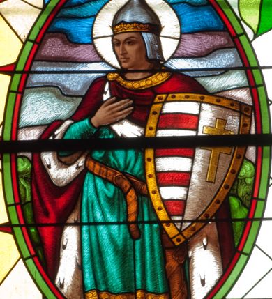 Zdjęcie nr 4: Witraż w kształcie stojącego prostokąta zamkniętego łukiem półkolistym. Kompozycja ujęta geometryczną bordiurą w kolorach czerwonym oraz jasno- i ciemnożółtym. Wewnątrz pole w kształcie stojącego prostokąta zamkniętego łukiem półkolistym z wpisanymi dwoma ostrołukami tworzącymi rodzaj gotyckiego biforium. W jego górnej części motyw kłosów oraz winnych gron. W polu z lewej strony kompozycji owalny medalion mieszczący przedstawienie św. Władysława w rycerskiej zbroi, zielonej tunice ze złotymi lamówkami i królewskim, czerwonym płaszczu z gronostajowym podbiciem umieszczone na tle pejzażu. Święty ukazany w tzw. długim popiersiu z ciałem zwróconym w trzech czwartych w prawo. Prawa ręka złożona na piersi, w lewej tarcza podzielona w słup, po prawej stronie białe i czerwone pasy, po lewej na srebrnym polu złoty krzyż z dwiema belkami. Na głowie hełm z koroną, wokół głowy nimb. Poniżej medalionu czerwona litera „A” w jasnożółtym polu w kształcie rombu oraz inskrypcja: „FUNDACJA / KS(IĄDZ) WŁADYSŁAW ZIEMIŃSKI PROBOSZCZ / R(OKU) P(AŃSKIEGO) 1937”. W polu z prawej strony kompozycji owalny medalion mieszczący scenę Nauczania Marii przez św. Annę umieszczoną na tle pejzażu. Święta Anna ukazana w pozycji siedzącej, z ciałem zwróconym w trzech czwartych w prawo, pochyla się ku stojącej po jej prawej stronie Marii. Lewa dłoń uniesiona w geście nauczania. Anna przedstawiona jako dojrzała kobieta ubrana w zieloną suknię, białą podwikę oraz czerwony płaszcz. Na głowie jasnozielona chusta. Maria ukazana z profilu w pozycji stojącej. Została przedstawiona jako kilkuletnia dziewczyna o długich, brązowych i falowanych włosach ujętych jasną opaską. Wokół głowy nimb. Ręce złożone w geście modlitwy. Postać ubrana jest w jasnoniebieską tunikę. Poniżej medaliony litera „Ω” w jasnożółtym polu w kształcie rombu oraz inskrypcja „FRANASOWA ANNA R(OKU) P(AŃSKIEGO) 1937”. 

