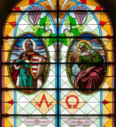 Zdjęcie nr 1: Witraż w kształcie stojącego prostokąta zamkniętego łukiem półkolistym. Kompozycja ujęta geometryczną bordiurą w kolorach czerwonym oraz jasno- i ciemnożółtym. Wewnątrz pole w kształcie stojącego prostokąta zamkniętego łukiem półkolistym z wpisanymi dwoma ostrołukami tworzącymi rodzaj gotyckiego biforium. W jego górnej części motyw kłosów oraz winnych gron. W polu z lewej strony kompozycji owalny medalion mieszczący przedstawienie św. Władysława w rycerskiej zbroi, zielonej tunice ze złotymi lamówkami i królewskim, czerwonym płaszczu z gronostajowym podbiciem umieszczone na tle pejzażu. Święty ukazany w tzw. długim popiersiu z ciałem zwróconym w trzech czwartych w prawo. Prawa ręka złożona na piersi, w lewej tarcza podzielona w słup, po prawej stronie białe i czerwone pasy, po lewej na srebrnym polu złoty krzyż z dwiema belkami. Na głowie hełm z koroną, wokół głowy nimb. Poniżej medalionu czerwona litera „A” w jasnożółtym polu w kształcie rombu oraz inskrypcja: „FUNDACJA / KS(IĄDZ) WŁADYSŁAW ZIEMIŃSKI PROBOSZCZ / R(OKU) P(AŃSKIEGO) 1937”. W polu z prawej strony kompozycji owalny medalion mieszczący scenę Nauczania Marii przez św. Annę umieszczoną na tle pejzażu. Święta Anna ukazana w pozycji siedzącej, z ciałem zwróconym w trzech czwartych w prawo, pochyla się ku stojącej po jej prawej stronie Marii. Lewa dłoń uniesiona w geście nauczania. Anna przedstawiona jako dojrzała kobieta ubrana w zieloną suknię, białą podwikę oraz czerwony płaszcz. Na głowie jasnozielona chusta. Maria ukazana z profilu w pozycji stojącej. Została przedstawiona jako kilkuletnia dziewczyna o długich, brązowych i falowanych włosach ujętych jasną opaską. Wokół głowy nimb. Ręce złożone w geście modlitwy. Postać ubrana jest w jasnoniebieską tunikę. Poniżej medaliony litera „Ω” w jasnożółtym polu w kształcie rombu oraz inskrypcja „FRANASOWA ANNA R(OKU) P(AŃSKIEGO) 1937”. 

