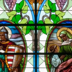 Zdjęcie nr 1: Witraż w kształcie stojącego prostokąta zamkniętego łukiem półkolistym. Kompozycja ujęta geometryczną bordiurą w kolorach czerwonym oraz jasno- i ciemnożółtym. Wewnątrz pole w kształcie stojącego prostokąta zamkniętego łukiem półkolistym z wpisanymi dwoma ostrołukami tworzącymi rodzaj gotyckiego biforium. W jego górnej części motyw kłosów oraz winnych gron. W polu z lewej strony kompozycji owalny medalion mieszczący przedstawienie św. Władysława w rycerskiej zbroi, zielonej tunice ze złotymi lamówkami i królewskim, czerwonym płaszczu z gronostajowym podbiciem umieszczone na tle pejzażu. Święty ukazany w tzw. długim popiersiu z ciałem zwróconym w trzech czwartych w prawo. Prawa ręka złożona na piersi, w lewej tarcza podzielona w słup, po prawej stronie białe i czerwone pasy, po lewej na srebrnym polu złoty krzyż z dwiema belkami. Na głowie hełm z koroną, wokół głowy nimb. Poniżej medalionu czerwona litera „A” w jasnożółtym polu w kształcie rombu oraz inskrypcja: „FUNDACJA / KS(IĄDZ) WŁADYSŁAW ZIEMIŃSKI PROBOSZCZ / R(OKU) P(AŃSKIEGO) 1937”. W polu z prawej strony kompozycji owalny medalion mieszczący scenę Nauczania Marii przez św. Annę umieszczoną na tle pejzażu. Święta Anna ukazana w pozycji siedzącej, z ciałem zwróconym w trzech czwartych w prawo, pochyla się ku stojącej po jej prawej stronie Marii. Lewa dłoń uniesiona w geście nauczania. Anna przedstawiona jako dojrzała kobieta ubrana w zieloną suknię, białą podwikę oraz czerwony płaszcz. Na głowie jasnozielona chusta. Maria ukazana z profilu w pozycji stojącej. Została przedstawiona jako kilkuletnia dziewczyna o długich, brązowych i falowanych włosach ujętych jasną opaską. Wokół głowy nimb. Ręce złożone w geście modlitwy. Postać ubrana jest w jasnoniebieską tunikę. Poniżej medaliony litera „Ω” w jasnożółtym polu w kształcie rombu oraz inskrypcja „FRANASOWA ANNA R(OKU) P(AŃSKIEGO) 1937”. 

