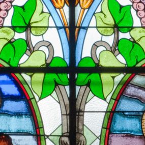 Zdjęcie nr 1: Witraż w kształcie stojącego prostokąta zamkniętego łukiem półkolistym. Kompozycja ujęta geometryczną bordiurą w kolorach czerwonym oraz jasno- i ciemnożółtym. Wewnątrz pole w kształcie stojącego prostokąta zamkniętego łukiem półkolistym z wpisanymi dwoma ostrołukami tworzącymi rodzaj gotyckiego biforium. W jego górnej części motyw kłosów oraz winnych gron. W polu z lewej strony kompozycji owalny medalion mieszczący przedstawienie św. Franciszka z Asyżu ubranego w brązowy habit przepasany sznurem, z kapturem na głowie, umieszczone na tle pejzażu. Święty ukazany frontalnie w tzw. długim popiersiu. W prawej ręce trzyma podwójny krzyż, w lewej otwartą księgę. Wokół głowy nimb. Poniżej medalionu godło tercjarzy (skrzyżowane ręce Chrystusa i św. Franciszka ze śladami po gwoździach w dłoniach, między którymi złoty krzyż na przecięciu ramion z koroną cierniową, u dołu błękitna wstęga) w jasnozielonym polu w kształcie rombu oraz inskrypcja „FUNDACJA / III ZAKON TERCJARZY R(OKU) P(AŃSKIEGO) 1937”. W polu z prawej strony kompozycji owalny medalion mieszczący wizerunek Matki Boskiej Śnieżnej na tle błękitnego nieba. Maria ukazana frontalnie, w tzw. długim popiersiu z głową zwróconą w trzech czwartych w lewo. Lewą ręką podtrzymuje Dzieciątko, prawa dłoń na tej samej wysokości z wyprostowanym palcem wskazującym i środkowym. Maria przedstawiona jako kobieta o owalnej twarzy z dużymi szeroko otwartymi oczami, prostym nosem i niewielkich, wąskich ustach. Jest ubrana w złote szaty – suknię z długimi rękawami oraz bogato zdobiony ornamentem roślinnym płaszcz. Na głowie złota chusta zdobiona ornamentem roślinnym oraz rozbudowana korona w typie zamkniętym zwieńczona krzyżem. Wokół głowy promienisty nimb. Dzieciątko przedstawione w pozycji siedzącej z ciałem zwróconym w trzech czwartych w kierunku Marii. Głowa uniesiona lekko do góry, prawa dłoń wyciągnięta do przodu wykonuje gest błogosławieństwa. W lewej dłoni Dzieciątko trzyma zamkniętą księgę. Postać jest ubrana w złotą tunikę z długimi rękawami zdobioną ornamentem roślinnym. Spod szat widoczne bose stopy. Na głowie Dzieciątka korona w typie zamkniętym zwieńczona krzyżem. Poniżej inskrypcja „FUNDACJA / RÓŻE MĘŻCZYZN I KOBIET R(OKU) P(AŃSKIEGO) 1937” oraz w prawym, dolnym rogu kompozycji sygnatura „Wy[konał] M[aksymilian] Romańczyk / Kraków”. Tło witraża wypełnia ukośna kratownica w kolorach jasnozielonym i białym (szkło bezbarwne).



