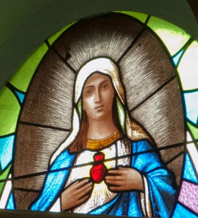 Zdjęcie nr 1: Witraż w kształcie wydłużonego owalu. W centrum kompozycji, na geometrycznym tle owalny medalion mieszczący półpostaciowe, frontalne przedstawienie Matki Boskiej wskazującej na serce widoczne na jej piersi. Maria ukazana jako młoda kobieta o owalnej twarzy z długim, prostym nosem i niewielkimi ustami. Oczy skierowane na wprost. Obie dłonie na wysokości piersi, wskazujące na serce umieszczone na tle glorii promienistej. Matka Boska ubrana w białą suknię z długimi rękawami wykończoną pod szyją szeroką, złotą lamówką oraz błękitny płaszcz. Na głowie Marii biała chusta o jasnozielonej podszewce. Wokół postaci gloria promienista.


