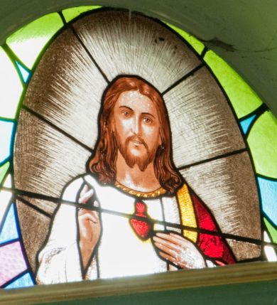 Zdjęcie nr 1: Witraż w kształcie wydłużonego owalu. W centrum kompozycji, na geometrycznym tle owalny medalion mieszczący półpostaciowe, frontalne przedstawienie Najświętszego Serca Jezusa. Chrystus ukazany jako młody mężczyzna o owalnej twarzy z długim, prostym nosem i niewielkimi ustami. Długie, jasnobrązowe włosy opadają na ramiona postaci. Krótka broda rozdwaja się na końcu. Oczy postaci skierowane na wprost. Chrystus lewą dłonią wskazuje na widoczne na piersi gorejące serce umieszczone na tle glorii promienistej,  prawą wykonuje gest błogosławieństwa. Ubrany jest w białą szatę z długimi rękawami wykończoną pod szyją szeroką, złotą lamówką oraz czerwono-złoty płaszcz luźno zarzucony na lewe ramię. Wokół postaci gloria promienista.

