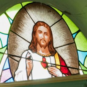 Zdjęcie nr 1: Witraż w kształcie wydłużonego owalu. W centrum kompozycji, na geometrycznym tle owalny medalion mieszczący półpostaciowe, frontalne przedstawienie Najświętszego Serca Jezusa. Chrystus ukazany jako młody mężczyzna o owalnej twarzy z długim, prostym nosem i niewielkimi ustami. Długie, jasnobrązowe włosy opadają na ramiona postaci. Krótka broda rozdwaja się na końcu. Oczy postaci skierowane na wprost. Chrystus lewą dłonią wskazuje na widoczne na piersi gorejące serce umieszczone na tle glorii promienistej,  prawą wykonuje gest błogosławieństwa. Ubrany jest w białą szatę z długimi rękawami wykończoną pod szyją szeroką, złotą lamówką oraz czerwono-złoty płaszcz luźno zarzucony na lewe ramię. Wokół postaci gloria promienista.

