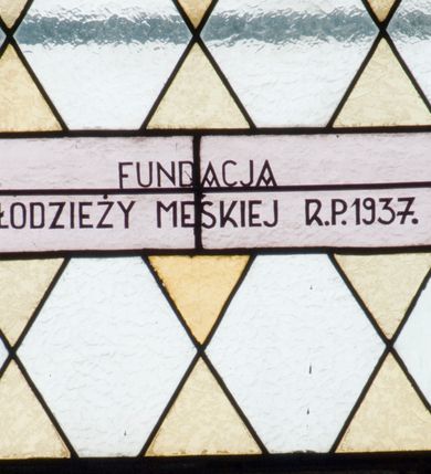 Zdjęcie nr 4: Witraż w kształcie wydłużonego, stojącego prostokąta zamkniętego półkoliście. W centrum kompozycji medalion mieszczący popiersie św. Stanisława Kostki. Święty zwrócony w trzech czwartych w prawo. Twarz owalna, o regularnych rysach. Ręce na wysokości piersi złożone w geście modlitwy. Poniżej bukiet białych lilii. Święty Stanisław ubrany jest w czarną sutannę z białym kołnierzem. Wokół głowy jasnożółty nimb. W górnej części witrażu przedstawione winne grona na tle geometrycznej kompozycji w kolorach żółtym i czerwonym. W dolnej strefie witrażu inskrypcja „FUNDACJA / MŁODZIEŻY MĘSKIEJ R(OKU) P(AŃSKIEGO) 1937”. Tło kompozycji wypełnione motywem rombów w kolorach jasnożółtym i białym.

