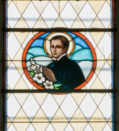 Zdjęcie nr 1: Witraż w kształcie wydłużonego, stojącego prostokąta zamkniętego półkoliście. W centrum kompozycji medalion mieszczący popiersie św. Stanisława Kostki. Święty zwrócony w trzech czwartych w prawo. Twarz owalna, o regularnych rysach. Ręce na wysokości piersi złożone w geście modlitwy. Poniżej bukiet białych lilii. Święty Stanisław ubrany jest w czarną sutannę z białym kołnierzem. Wokół głowy jasnożółty nimb. W górnej części witrażu przedstawione winne grona na tle geometrycznej kompozycji w kolorach żółtym i czerwonym. W dolnej strefie witrażu inskrypcja „FUNDACJA / MŁODZIEŻY MĘSKIEJ R(OKU) P(AŃSKIEGO) 1937”. Tło kompozycji wypełnione motywem rombów w kolorach jasnożółtym i białym.

