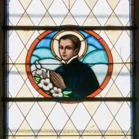 Zdjęcie nr 1: Witraż w kształcie wydłużonego, stojącego prostokąta zamkniętego półkoliście. W centrum kompozycji medalion mieszczący popiersie św. Stanisława Kostki. Święty zwrócony w trzech czwartych w prawo. Twarz owalna, o regularnych rysach. Ręce na wysokości piersi złożone w geście modlitwy. Poniżej bukiet białych lilii. Święty Stanisław ubrany jest w czarną sutannę z białym kołnierzem. Wokół głowy jasnożółty nimb. W górnej części witrażu przedstawione winne grona na tle geometrycznej kompozycji w kolorach żółtym i czerwonym. W dolnej strefie witrażu inskrypcja „FUNDACJA / MŁODZIEŻY MĘSKIEJ R(OKU) P(AŃSKIEGO) 1937”. Tło kompozycji wypełnione motywem rombów w kolorach jasnożółtym i białym.

