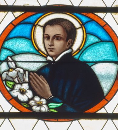 Zdjęcie nr 2: Witraż w kształcie wydłużonego, stojącego prostokąta zamkniętego półkoliście. W centrum kompozycji medalion mieszczący popiersie św. Stanisława Kostki. Święty zwrócony w trzech czwartych w prawo. Twarz owalna, o regularnych rysach. Ręce na wysokości piersi złożone w geście modlitwy. Poniżej bukiet białych lilii. Święty Stanisław ubrany jest w czarną sutannę z białym kołnierzem. Wokół głowy jasnożółty nimb. W górnej części witrażu przedstawione winne grona na tle geometrycznej kompozycji w kolorach żółtym i czerwonym. W dolnej strefie witrażu inskrypcja „FUNDACJA / MŁODZIEŻY MĘSKIEJ R(OKU) P(AŃSKIEGO) 1937”. Tło kompozycji wypełnione motywem rombów w kolorach jasnożółtym i białym.

