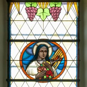 Zdjęcie nr 1: Witraż w kształcie wydłużonego, stojącego prostokąta zamkniętego półkoliście. W centrum kompozycji umieszczony medalion mieszczący popiersie św. Teresy z Lisieux trzymającej krucyfiks i róże. Święta ukazana frontalnie z głową zwróconą w trzech czwartych w lewo. Twarz owalna, o regularnych rysach, z dużymi oczami, wąskim nosem oraz niewielkimi ustami. Święta w rękach złożonych na wysokości piersi trzyma bukiet czerwonych i różowych róż oraz krucyfiks, na który spływają promienie. Teresa ubrana jest w brązowy karmelitański habit z białym płaszczem oraz białą toką i czarnym welonem na głowie. Tło medalionu utrzymane jest odcieniach niebieskiego. Wokół głowy jasnożółty nimb. W górnej części witrażu przedstawione winne grona na tle geometrycznej kompozycji w kolorach żółtym i czerwonym. W dolnej strefie witrażu inskrypcja „FUNDACJA / MŁODZIEŻY ŻEŃSKIEJ R(OKU) P(AŃSKIEGO) 1937” oraz poniżej, w lewym, dolnym rogu sygnatura: „Wy[konał] / M[aksymilian]Romańczyk / Kraków św. Jana 30”. Tło kompozycji wypełnione motywem rombów w kolorach jasnożółtym i białym.