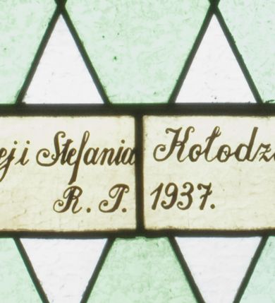 Zdjęcie nr 3: Witraż w kształcie stojącego prostokąta zamkniętego półkoliście ujęty geometryczną bordiurą dekorowaną czerwonymi i żółtymi prostokątami. W polu ukośna kratka wypełniona białymi i jasnozielonymi rombami. W górnej części witraża okrągły medalion z czerwonym monogramem IHS umieszczonym na żółtym tle. W dolej kwaterze prostokątne pole z napisem: „Fund[acja]: Maciej i Stefania Kołodziejowie / R[OKU] P[AŃSKIEGO] 1937&quot;.