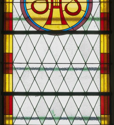 Zdjęcie nr 2: Witraż w kształcie stojącego prostokąta zamkniętego półkoliście ujęty geometryczną bordiurą dekorowaną czerwonymi i żółtymi prostokątami. W polu ukośna kratka wypełniona białymi i jasnozielonymi rombami. W górnej części witraża okrągły medalion z czerwonym monogramem IHS umieszczonym na żółtym tle. W dolej kwaterze prostokątne pole z napisem: „Fund[acja]: Maciej i Stefania Kołodziejowie / R[OKU] P[AŃSKIEGO] 1937&quot;.