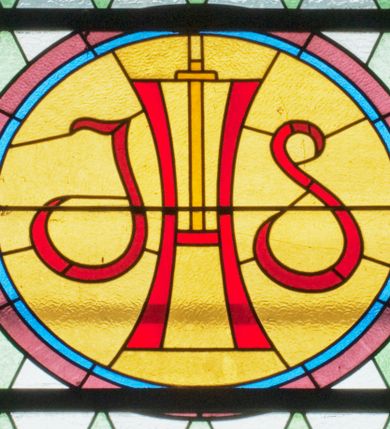 Zdjęcie nr 1: Witraż w kształcie stojącego prostokąta zamkniętego półkoliście ujęty geometryczną bordiurą dekorowaną czerwonymi i żółtymi prostokątami. W polu ukośna kratka wypełniona białymi i jasnozielonymi rombami. W górnej części witraża okrągły medalion z czerwonym monogramem IHS umieszczonym na żółtym tle. W dolej kwaterze prostokątne pole z napisem: „Fund[acja]: Maciej i Stefania Kołodziejowie / R[OKU] P[AŃSKIEGO] 1937&quot;.