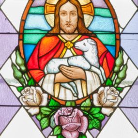 Zdjęcie nr 1: Witraż w kształcie stojącego prostokąta zamkniętego półkoliście ujęty geometryczną bordiurą wypełnioną błękitnymi i jasnozielonymi trójkątami oraz ciemnoczerwonymi rombami. W polu ukośna krata wypełniona naprzemiennie białymi i jasnofioletowymi rombami. W dolnej części witraża umieszczony owalny medalion wsparty na girlandzie z kwitnących róż mieszczący frontalne przedstawienie Chrystusa przedstawionego w typie Dobrego Pasterza. Chrystus ujęty w popiersiu, ubrany w białą szatę z długimi rękawami oraz czerwony płaszcz spięty pod szyją złotą klamrą w kształcie czwórliścia. Na rękach trzyma białego baranka. Wokół głowy postaci widoczny nimb krzyżowy. Tło medalionu w kolorach zielonym, fioletowym i odcieniach niebieskiego. Powyżej prostokątne pole z napisem: "Fund[ator]: Fr[anciszek] Grodowski i Józef Łosiński / inwalidzi wojenni / Wykonano R[oku] P[ańskiego] 1938 za pobytu Ks[iędza] Prob[oszcza] Wł[adysława] Ziemińskiego".