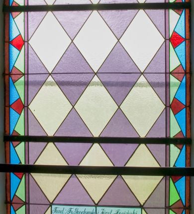 Zdjęcie nr 2: Witraż w kształcie stojącego prostokąta zamkniętego półkoliście ujęty geometryczną bordiurą wypełnioną błękitnymi i jasnozielonymi trójkątami oraz ciemnoczerwonymi rombami. W polu ukośna krata wypełniona naprzemiennie białymi i jasnofioletowymi rombami. W dolnej części witraża umieszczony owalny medalion wsparty na girlandzie z kwitnących róż mieszczący frontalne przedstawienie Chrystusa przedstawionego w typie Dobrego Pasterza. Chrystus ujęty w popiersiu, ubrany w białą szatę z długimi rękawami oraz czerwony płaszcz spięty pod szyją złotą klamrą w kształcie czwórliścia. Na rękach trzyma białego baranka. Wokół głowy postaci widoczny nimb krzyżowy. Tło medalionu w kolorach zielonym, fioletowym i odcieniach niebieskiego. Powyżej prostokątne pole z napisem: "Fund[ator]: Fr[anciszek] Grodowski i Józef Łosiński / inwalidzi wojenni / Wykonano R[oku] P[ańskiego] 1938 za pobytu Ks[iędza] Prob[oszcza] Wł[adysława] Ziemińskiego".