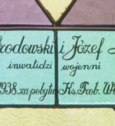 Zdjęcie nr 3: Witraż w kształcie stojącego prostokąta zamkniętego półkoliście ujęty geometryczną bordiurą wypełnioną błękitnymi i jasnozielonymi trójkątami oraz ciemnoczerwonymi rombami. W polu ukośna krata wypełniona naprzemiennie białymi i jasnofioletowymi rombami. W dolnej części witraża umieszczony owalny medalion wsparty na girlandzie z kwitnących róż mieszczący frontalne przedstawienie Chrystusa przedstawionego w typie Dobrego Pasterza. Chrystus ujęty w popiersiu, ubrany w białą szatę z długimi rękawami oraz czerwony płaszcz spięty pod szyją złotą klamrą w kształcie czwórliścia. Na rękach trzyma białego baranka. Wokół głowy postaci widoczny nimb krzyżowy. Tło medalionu w kolorach zielonym, fioletowym i odcieniach niebieskiego. Powyżej prostokątne pole z napisem: "Fund[ator]: Fr[anciszek] Grodowski i Józef Łosiński / inwalidzi wojenni / Wykonano R[oku] P[ańskiego] 1938 za pobytu Ks[iędza] Prob[oszcza] Wł[adysława] Ziemińskiego".