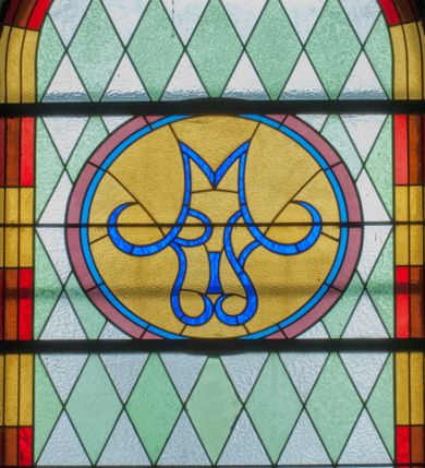 Zdjęcie nr 1: Witraż w kształcie stojącego prostokąta zamkniętego półkoliście ujęty geometryczną bordiurą dekorowaną czerwonymi i żółtymi prostokątami. W polu ukośna kratka wypełniona białymi i jasnozielonymi rombami. W górnej części witraża okrągły medalion z niebieskim monogamem maryjnym umieszczonym na żółtym tle. W dolej kwaterze prostokątne pole z napisem: „FUNDACJA / MAGDALENA HARDENOWA R[OKU] P[AŃSKIEGO] 1937.” 