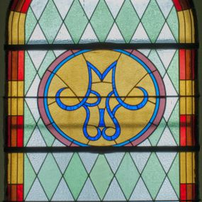 Zdjęcie nr 1: Witraż w kształcie stojącego prostokąta zamkniętego półkoliście ujęty geometryczną bordiurą dekorowaną czerwonymi i żółtymi prostokątami. W polu ukośna kratka wypełniona białymi i jasnozielonymi rombami. W górnej części witraża okrągły medalion z niebieskim monogamem maryjnym umieszczonym na żółtym tle. W dolej kwaterze prostokątne pole z napisem: „FUNDACJA / MAGDALENA HARDENOWA R[OKU] P[AŃSKIEGO] 1937.” 