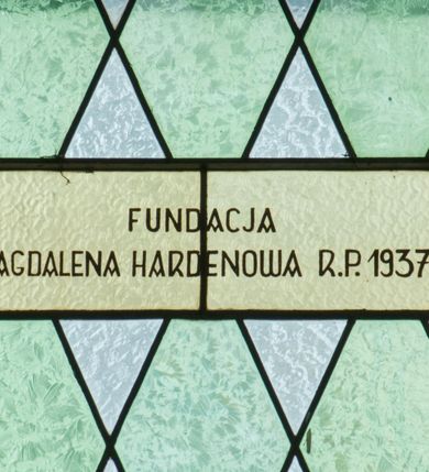 Zdjęcie nr 3: Witraż w kształcie stojącego prostokąta zamkniętego półkoliście ujęty geometryczną bordiurą dekorowaną czerwonymi i żółtymi prostokątami. W polu ukośna kratka wypełniona białymi i jasnozielonymi rombami. W górnej części witraża okrągły medalion z niebieskim monogamem maryjnym umieszczonym na żółtym tle. W dolej kwaterze prostokątne pole z napisem: „FUNDACJA / MAGDALENA HARDENOWA R[OKU] P[AŃSKIEGO] 1937.” 