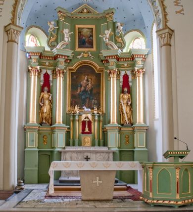 Zdjęcie nr 1: Ołtarz przyścienny, architektoniczny, trójosiowy, dwukondygnacyjny ze zwieńczeniem. Nastawa umieszczona na wysokim, dwudzielnym cokole w górnej części zdobionym na przemian dekoracją płycinową i motywem muszli palmetowej z podwieszoną girlandą kwiatową oraz rozetkami. Cokół wieńczy profilowany gzyms dekorowany kostkowaniem. Antepedium współczesne, marmurowe z umieszczonym centralnie prostym, równoramiennym krzyżem. Na mensie ustawione tabernakulum z tronem wystawienia, o formach architektonicznych, ujęte zdwojonymi, smukłymi kolumienkami o złoconych trzonach umieszczonymi na wysokich cokołach. Podpory dźwigają odcinki pełnego belkowania, na których ustawione złocone figury klęczących aniołów. Część centralna o linii wypukłej, dwukondygnacyjna. W kondygnacji pierwszej złocone drzwiczki z płaskorzeźbionym kielichem z hostią na tle glorii promienistej, powyżej, w kondygnacji drugiej tron wystawienia ujęty rzeźbioną, złoconą kotarą. W zwieńczeniu srebrzona figura baranka apokaliptycznego leżącego na księdze. Nastawa ujęta zdwojonymi, kompozytowymi kolumnami o złoconych trzonach i kapitelach. Pomiędzy kolumnami, w niszach rzeźbione figury św. Piotra (z lewej) i św. Pawła (z prawej), ponad którymi motyw symetrycznie podwieszonej kotary. Kolumny dźwigają odcinki pełnego belkowania z fryzem dekorowanym rozetkami i uskrzydlonymi główkami anielskimi oraz wydatnym gzymsem dekorowanym kostkowaniem. Na osi wewnętrznej pary komun dodatkowo ustawione odcinki półkolistego tympanonu, na których siedzą putta. W polu głównym ołtarza złocona, profilowana rama w kształcie stojącego prostokąta zamkniętego łukiem nadwieszonym mieszcząca barokowy obraz Matki Boskiej z Dzieciątkiem. Nieco powyżej, w osi ołtarza dwa putta unoszące złoconą koronę typu zamkniętego zwieńczoną krzyżem. Kondygnacja druga ujęta parą pilastrów o kompozytowych kapitelach oraz uszami w formie spływów wolutowych, na których siedzące putta. W polu złocona i profilowana rama w kształcie stojącego prostokąta mieszcząca obraz przedstawiający Trójcę Świętą. Całość ołtarza wieńczy profilowany gzyms dekorowany kostkowaniem i zamknięty niskim, trójkątnym przyczółkiem. W polu tympanonu uskrzydlona główka anielska. Struktura ołtarza polichromowana w kolorze jasnozielonym z elementami czerwieni i bieli. Detale architektoniczne, ornamenty oraz szaty postaci złocone.

