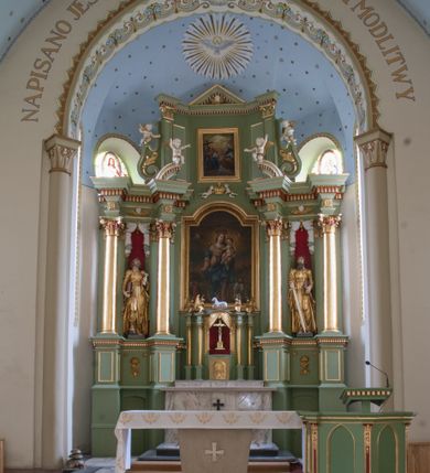 Zdjęcie nr 3: Ołtarz przyścienny, architektoniczny, trójosiowy, dwukondygnacyjny ze zwieńczeniem. Nastawa umieszczona na wysokim, dwudzielnym cokole w górnej części zdobionym na przemian dekoracją płycinową i motywem muszli palmetowej z podwieszoną girlandą kwiatową oraz rozetkami. Cokół wieńczy profilowany gzyms dekorowany kostkowaniem. Antepedium współczesne, marmurowe z umieszczonym centralnie prostym, równoramiennym krzyżem. Na mensie ustawione tabernakulum z tronem wystawienia, o formach architektonicznych, ujęte zdwojonymi, smukłymi kolumienkami o złoconych trzonach umieszczonymi na wysokich cokołach. Podpory dźwigają odcinki pełnego belkowania, na których ustawione złocone figury klęczących aniołów. Część centralna o linii wypukłej, dwukondygnacyjna. W kondygnacji pierwszej złocone drzwiczki z płaskorzeźbionym kielichem z hostią na tle glorii promienistej, powyżej, w kondygnacji drugiej tron wystawienia ujęty rzeźbioną, złoconą kotarą. W zwieńczeniu srebrzona figura baranka apokaliptycznego leżącego na księdze. Nastawa ujęta zdwojonymi, kompozytowymi kolumnami o złoconych trzonach i kapitelach. Pomiędzy kolumnami, w niszach rzeźbione figury św. Piotra (z lewej) i św. Pawła (z prawej), ponad którymi motyw symetrycznie podwieszonej kotary. Kolumny dźwigają odcinki pełnego belkowania z fryzem dekorowanym rozetkami i uskrzydlonymi główkami anielskimi oraz wydatnym gzymsem dekorowanym kostkowaniem. Na osi wewnętrznej pary komun dodatkowo ustawione odcinki półkolistego tympanonu, na których siedzą putta. W polu głównym ołtarza złocona, profilowana rama w kształcie stojącego prostokąta zamkniętego łukiem nadwieszonym mieszcząca barokowy obraz Matki Boskiej z Dzieciątkiem. Nieco powyżej, w osi ołtarza dwa putta unoszące złoconą koronę typu zamkniętego zwieńczoną krzyżem. Kondygnacja druga ujęta parą pilastrów o kompozytowych kapitelach oraz uszami w formie spływów wolutowych, na których siedzące putta. W polu złocona i profilowana rama w kształcie stojącego prostokąta mieszcząca obraz przedstawiający Trójcę Świętą. Całość ołtarza wieńczy profilowany gzyms dekorowany kostkowaniem i zamknięty niskim, trójkątnym przyczółkiem. W polu tympanonu uskrzydlona główka anielska. Struktura ołtarza polichromowana w kolorze jasnozielonym z elementami czerwieni i bieli. Detale architektoniczne, ornamenty oraz szaty postaci złocone.

