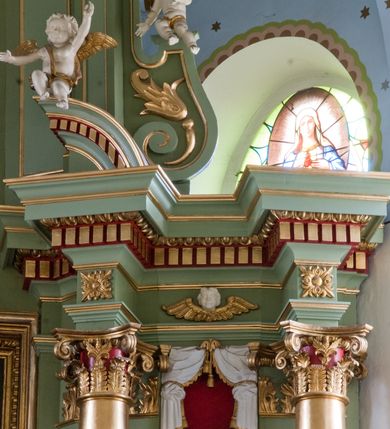 Zdjęcie nr 12: Ołtarz przyścienny, architektoniczny, trójosiowy, dwukondygnacyjny ze zwieńczeniem. Nastawa umieszczona na wysokim, dwudzielnym cokole w górnej części zdobionym na przemian dekoracją płycinową i motywem muszli palmetowej z podwieszoną girlandą kwiatową oraz rozetkami. Cokół wieńczy profilowany gzyms dekorowany kostkowaniem. Antepedium współczesne, marmurowe z umieszczonym centralnie prostym, równoramiennym krzyżem. Na mensie ustawione tabernakulum z tronem wystawienia, o formach architektonicznych, ujęte zdwojonymi, smukłymi kolumienkami o złoconych trzonach umieszczonymi na wysokich cokołach. Podpory dźwigają odcinki pełnego belkowania, na których ustawione złocone figury klęczących aniołów. Część centralna o linii wypukłej, dwukondygnacyjna. W kondygnacji pierwszej złocone drzwiczki z płaskorzeźbionym kielichem z hostią na tle glorii promienistej, powyżej, w kondygnacji drugiej tron wystawienia ujęty rzeźbioną, złoconą kotarą. W zwieńczeniu srebrzona figura baranka apokaliptycznego leżącego na księdze. Nastawa ujęta zdwojonymi, kompozytowymi kolumnami o złoconych trzonach i kapitelach. Pomiędzy kolumnami, w niszach rzeźbione figury św. Piotra (z lewej) i św. Pawła (z prawej), ponad którymi motyw symetrycznie podwieszonej kotary. Kolumny dźwigają odcinki pełnego belkowania z fryzem dekorowanym rozetkami i uskrzydlonymi główkami anielskimi oraz wydatnym gzymsem dekorowanym kostkowaniem. Na osi wewnętrznej pary komun dodatkowo ustawione odcinki półkolistego tympanonu, na których siedzą putta. W polu głównym ołtarza złocona, profilowana rama w kształcie stojącego prostokąta zamkniętego łukiem nadwieszonym mieszcząca barokowy obraz Matki Boskiej z Dzieciątkiem. Nieco powyżej, w osi ołtarza dwa putta unoszące złoconą koronę typu zamkniętego zwieńczoną krzyżem. Kondygnacja druga ujęta parą pilastrów o kompozytowych kapitelach oraz uszami w formie spływów wolutowych, na których siedzące putta. W polu złocona i profilowana rama w kształcie stojącego prostokąta mieszcząca obraz przedstawiający Trójcę Świętą. Całość ołtarza wieńczy profilowany gzyms dekorowany kostkowaniem i zamknięty niskim, trójkątnym przyczółkiem. W polu tympanonu uskrzydlona główka anielska. Struktura ołtarza polichromowana w kolorze jasnozielonym z elementami czerwieni i bieli. Detale architektoniczne, ornamenty oraz szaty postaci złocone.

