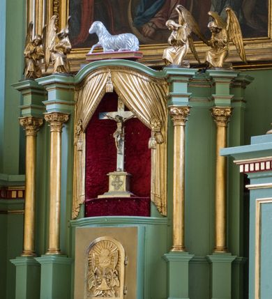Zdjęcie nr 14: Ołtarz przyścienny, architektoniczny, trójosiowy, dwukondygnacyjny ze zwieńczeniem. Nastawa umieszczona na wysokim, dwudzielnym cokole w górnej części zdobionym na przemian dekoracją płycinową i motywem muszli palmetowej z podwieszoną girlandą kwiatową oraz rozetkami. Cokół wieńczy profilowany gzyms dekorowany kostkowaniem. Antepedium współczesne, marmurowe z umieszczonym centralnie prostym, równoramiennym krzyżem. Na mensie ustawione tabernakulum z tronem wystawienia, o formach architektonicznych, ujęte zdwojonymi, smukłymi kolumienkami o złoconych trzonach umieszczonymi na wysokich cokołach. Podpory dźwigają odcinki pełnego belkowania, na których ustawione złocone figury klęczących aniołów. Część centralna o linii wypukłej, dwukondygnacyjna. W kondygnacji pierwszej złocone drzwiczki z płaskorzeźbionym kielichem z hostią na tle glorii promienistej, powyżej, w kondygnacji drugiej tron wystawienia ujęty rzeźbioną, złoconą kotarą. W zwieńczeniu srebrzona figura baranka apokaliptycznego leżącego na księdze. Nastawa ujęta zdwojonymi, kompozytowymi kolumnami o złoconych trzonach i kapitelach. Pomiędzy kolumnami, w niszach rzeźbione figury św. Piotra (z lewej) i św. Pawła (z prawej), ponad którymi motyw symetrycznie podwieszonej kotary. Kolumny dźwigają odcinki pełnego belkowania z fryzem dekorowanym rozetkami i uskrzydlonymi główkami anielskimi oraz wydatnym gzymsem dekorowanym kostkowaniem. Na osi wewnętrznej pary komun dodatkowo ustawione odcinki półkolistego tympanonu, na których siedzą putta. W polu głównym ołtarza złocona, profilowana rama w kształcie stojącego prostokąta zamkniętego łukiem nadwieszonym mieszcząca barokowy obraz Matki Boskiej z Dzieciątkiem. Nieco powyżej, w osi ołtarza dwa putta unoszące złoconą koronę typu zamkniętego zwieńczoną krzyżem. Kondygnacja druga ujęta parą pilastrów o kompozytowych kapitelach oraz uszami w formie spływów wolutowych, na których siedzące putta. W polu złocona i profilowana rama w kształcie stojącego prostokąta mieszcząca obraz przedstawiający Trójcę Świętą. Całość ołtarza wieńczy profilowany gzyms dekorowany kostkowaniem i zamknięty niskim, trójkątnym przyczółkiem. W polu tympanonu uskrzydlona główka anielska. Struktura ołtarza polichromowana w kolorze jasnozielonym z elementami czerwieni i bieli. Detale architektoniczne, ornamenty oraz szaty postaci złocone.

