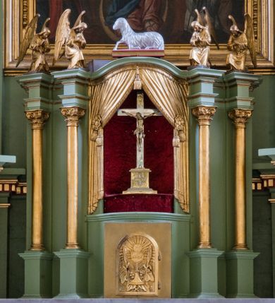 Zdjęcie nr 27: Ołtarz przyścienny, architektoniczny, trójosiowy, dwukondygnacyjny ze zwieńczeniem. Nastawa umieszczona na wysokim, dwudzielnym cokole w górnej części zdobionym na przemian dekoracją płycinową i motywem muszli palmetowej z podwieszoną girlandą kwiatową oraz rozetkami. Cokół wieńczy profilowany gzyms dekorowany kostkowaniem. Antepedium współczesne, marmurowe z umieszczonym centralnie prostym, równoramiennym krzyżem. Na mensie ustawione tabernakulum z tronem wystawienia, o formach architektonicznych, ujęte zdwojonymi, smukłymi kolumienkami o złoconych trzonach umieszczonymi na wysokich cokołach. Podpory dźwigają odcinki pełnego belkowania, na których ustawione złocone figury klęczących aniołów. Część centralna o linii wypukłej, dwukondygnacyjna. W kondygnacji pierwszej złocone drzwiczki z płaskorzeźbionym kielichem z hostią na tle glorii promienistej, powyżej, w kondygnacji drugiej tron wystawienia ujęty rzeźbioną, złoconą kotarą. W zwieńczeniu srebrzona figura baranka apokaliptycznego leżącego na księdze. Nastawa ujęta zdwojonymi, kompozytowymi kolumnami o złoconych trzonach i kapitelach. Pomiędzy kolumnami, w niszach rzeźbione figury św. Piotra (z lewej) i św. Pawła (z prawej), ponad którymi motyw symetrycznie podwieszonej kotary. Kolumny dźwigają odcinki pełnego belkowania z fryzem dekorowanym rozetkami i uskrzydlonymi główkami anielskimi oraz wydatnym gzymsem dekorowanym kostkowaniem. Na osi wewnętrznej pary komun dodatkowo ustawione odcinki półkolistego tympanonu, na których siedzą putta. W polu głównym ołtarza złocona, profilowana rama w kształcie stojącego prostokąta zamkniętego łukiem nadwieszonym mieszcząca barokowy obraz Matki Boskiej z Dzieciątkiem. Nieco powyżej, w osi ołtarza dwa putta unoszące złoconą koronę typu zamkniętego zwieńczoną krzyżem. Kondygnacja druga ujęta parą pilastrów o kompozytowych kapitelach oraz uszami w formie spływów wolutowych, na których siedzące putta. W polu złocona i profilowana rama w kształcie stojącego prostokąta mieszcząca obraz przedstawiający Trójcę Świętą. Całość ołtarza wieńczy profilowany gzyms dekorowany kostkowaniem i zamknięty niskim, trójkątnym przyczółkiem. W polu tympanonu uskrzydlona główka anielska. Struktura ołtarza polichromowana w kolorze jasnozielonym z elementami czerwieni i bieli. Detale architektoniczne, ornamenty oraz szaty postaci złocone.

