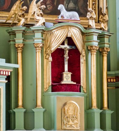 Zdjęcie nr 28: Ołtarz przyścienny, architektoniczny, trójosiowy, dwukondygnacyjny ze zwieńczeniem. Nastawa umieszczona na wysokim, dwudzielnym cokole w górnej części zdobionym na przemian dekoracją płycinową i motywem muszli palmetowej z podwieszoną girlandą kwiatową oraz rozetkami. Cokół wieńczy profilowany gzyms dekorowany kostkowaniem. Antepedium współczesne, marmurowe z umieszczonym centralnie prostym, równoramiennym krzyżem. Na mensie ustawione tabernakulum z tronem wystawienia, o formach architektonicznych, ujęte zdwojonymi, smukłymi kolumienkami o złoconych trzonach umieszczonymi na wysokich cokołach. Podpory dźwigają odcinki pełnego belkowania, na których ustawione złocone figury klęczących aniołów. Część centralna o linii wypukłej, dwukondygnacyjna. W kondygnacji pierwszej złocone drzwiczki z płaskorzeźbionym kielichem z hostią na tle glorii promienistej, powyżej, w kondygnacji drugiej tron wystawienia ujęty rzeźbioną, złoconą kotarą. W zwieńczeniu srebrzona figura baranka apokaliptycznego leżącego na księdze. Nastawa ujęta zdwojonymi, kompozytowymi kolumnami o złoconych trzonach i kapitelach. Pomiędzy kolumnami, w niszach rzeźbione figury św. Piotra (z lewej) i św. Pawła (z prawej), ponad którymi motyw symetrycznie podwieszonej kotary. Kolumny dźwigają odcinki pełnego belkowania z fryzem dekorowanym rozetkami i uskrzydlonymi główkami anielskimi oraz wydatnym gzymsem dekorowanym kostkowaniem. Na osi wewnętrznej pary komun dodatkowo ustawione odcinki półkolistego tympanonu, na których siedzą putta. W polu głównym ołtarza złocona, profilowana rama w kształcie stojącego prostokąta zamkniętego łukiem nadwieszonym mieszcząca barokowy obraz Matki Boskiej z Dzieciątkiem. Nieco powyżej, w osi ołtarza dwa putta unoszące złoconą koronę typu zamkniętego zwieńczoną krzyżem. Kondygnacja druga ujęta parą pilastrów o kompozytowych kapitelach oraz uszami w formie spływów wolutowych, na których siedzące putta. W polu złocona i profilowana rama w kształcie stojącego prostokąta mieszcząca obraz przedstawiający Trójcę Świętą. Całość ołtarza wieńczy profilowany gzyms dekorowany kostkowaniem i zamknięty niskim, trójkątnym przyczółkiem. W polu tympanonu uskrzydlona główka anielska. Struktura ołtarza polichromowana w kolorze jasnozielonym z elementami czerwieni i bieli. Detale architektoniczne, ornamenty oraz szaty postaci złocone.

