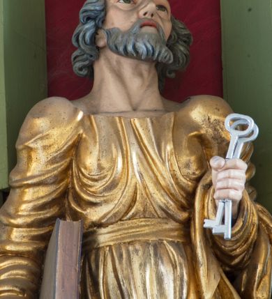 Zdjęcie nr 5: Rzeźba drążona ustawiona na niewielkim, prostopadłościennym postumencie. Święty Piotr został przedstawiony całopostaciowo, w pozycji stojącej z prawą nogą wspartą na kamieniu. W lewej ręce trzyma dwa srebrne klucze, na prawej, opuszczonej wzdłuż tułowia podtrzymuje księgę. Święty został przedstawiony jako starszy mężczyzna o wyrazistych rysach twarzy – głęboko osadzonych oczach, krótkim i wąskim nosie oraz wystających kościach policzkowych. Na głowie siwe, kręcone włosy przerzedzone ponad czołem oraz również siwy zarost. Wzrok postaci skierowany ku górze. Święty Piotr jest ubrany w złocone, bogato fałdowane szaty – sięgającą ziemi suknię z długimi, wąskimi rękawami przewiązaną w pasie oraz złoty płaszcz udrapowany wokół lewej ręki. Widoczne spod szat stopy postaci są bose. Figura polichromowana, partie szat złocone.

