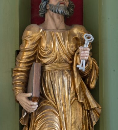 Zdjęcie nr 3: Rzeźba drążona ustawiona na niewielkim, prostopadłościennym postumencie. Święty Piotr został przedstawiony całopostaciowo, w pozycji stojącej z prawą nogą wspartą na kamieniu. W lewej ręce trzyma dwa srebrne klucze, na prawej, opuszczonej wzdłuż tułowia podtrzymuje księgę. Święty został przedstawiony jako starszy mężczyzna o wyrazistych rysach twarzy – głęboko osadzonych oczach, krótkim i wąskim nosie oraz wystających kościach policzkowych. Na głowie siwe, kręcone włosy przerzedzone ponad czołem oraz również siwy zarost. Wzrok postaci skierowany ku górze. Święty Piotr jest ubrany w złocone, bogato fałdowane szaty – sięgającą ziemi suknię z długimi, wąskimi rękawami przewiązaną w pasie oraz złoty płaszcz udrapowany wokół lewej ręki. Widoczne spod szat stopy postaci są bose. Figura polichromowana, partie szat złocone.

