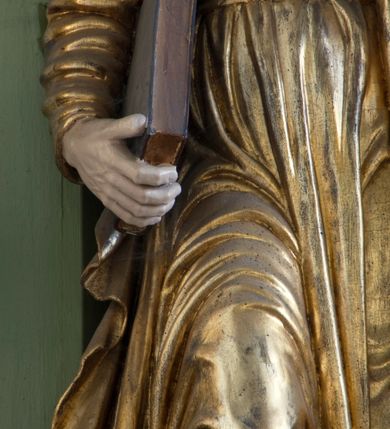 Zdjęcie nr 6: Rzeźba drążona ustawiona na niewielkim, prostopadłościennym postumencie. Święty Piotr został przedstawiony całopostaciowo, w pozycji stojącej z prawą nogą wspartą na kamieniu. W lewej ręce trzyma dwa srebrne klucze, na prawej, opuszczonej wzdłuż tułowia podtrzymuje księgę. Święty został przedstawiony jako starszy mężczyzna o wyrazistych rysach twarzy – głęboko osadzonych oczach, krótkim i wąskim nosie oraz wystających kościach policzkowych. Na głowie siwe, kręcone włosy przerzedzone ponad czołem oraz również siwy zarost. Wzrok postaci skierowany ku górze. Święty Piotr jest ubrany w złocone, bogato fałdowane szaty – sięgającą ziemi suknię z długimi, wąskimi rękawami przewiązaną w pasie oraz złoty płaszcz udrapowany wokół lewej ręki. Widoczne spod szat stopy postaci są bose. Figura polichromowana, partie szat złocone.

