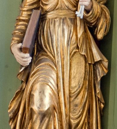 Zdjęcie nr 4: Rzeźba drążona ustawiona na niewielkim, prostopadłościennym postumencie. Święty Piotr został przedstawiony całopostaciowo, w pozycji stojącej z prawą nogą wspartą na kamieniu. W lewej ręce trzyma dwa srebrne klucze, na prawej, opuszczonej wzdłuż tułowia podtrzymuje księgę. Święty został przedstawiony jako starszy mężczyzna o wyrazistych rysach twarzy – głęboko osadzonych oczach, krótkim i wąskim nosie oraz wystających kościach policzkowych. Na głowie siwe, kręcone włosy przerzedzone ponad czołem oraz również siwy zarost. Wzrok postaci skierowany ku górze. Święty Piotr jest ubrany w złocone, bogato fałdowane szaty – sięgającą ziemi suknię z długimi, wąskimi rękawami przewiązaną w pasie oraz złoty płaszcz udrapowany wokół lewej ręki. Widoczne spod szat stopy postaci są bose. Figura polichromowana, partie szat złocone.

