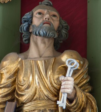 Zdjęcie nr 7: Rzeźba drążona ustawiona na niewielkim, prostopadłościennym postumencie. Święty Piotr został przedstawiony całopostaciowo, w pozycji stojącej z prawą nogą wspartą na kamieniu. W lewej ręce trzyma dwa srebrne klucze, na prawej, opuszczonej wzdłuż tułowia podtrzymuje księgę. Święty został przedstawiony jako starszy mężczyzna o wyrazistych rysach twarzy – głęboko osadzonych oczach, krótkim i wąskim nosie oraz wystających kościach policzkowych. Na głowie siwe, kręcone włosy przerzedzone ponad czołem oraz również siwy zarost. Wzrok postaci skierowany ku górze. Święty Piotr jest ubrany w złocone, bogato fałdowane szaty – sięgającą ziemi suknię z długimi, wąskimi rękawami przewiązaną w pasie oraz złoty płaszcz udrapowany wokół lewej ręki. Widoczne spod szat stopy postaci są bose. Figura polichromowana, partie szat złocone.

