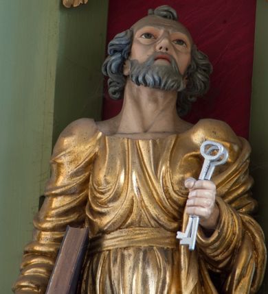 Zdjęcie nr 9: Rzeźba drążona ustawiona na niewielkim, prostopadłościennym postumencie. Święty Piotr został przedstawiony całopostaciowo, w pozycji stojącej z prawą nogą wspartą na kamieniu. W lewej ręce trzyma dwa srebrne klucze, na prawej, opuszczonej wzdłuż tułowia podtrzymuje księgę. Święty został przedstawiony jako starszy mężczyzna o wyrazistych rysach twarzy – głęboko osadzonych oczach, krótkim i wąskim nosie oraz wystających kościach policzkowych. Na głowie siwe, kręcone włosy przerzedzone ponad czołem oraz również siwy zarost. Wzrok postaci skierowany ku górze. Święty Piotr jest ubrany w złocone, bogato fałdowane szaty – sięgającą ziemi suknię z długimi, wąskimi rękawami przewiązaną w pasie oraz złoty płaszcz udrapowany wokół lewej ręki. Widoczne spod szat stopy postaci są bose. Figura polichromowana, partie szat złocone.

