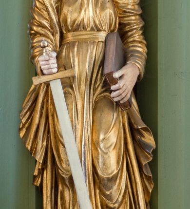 Zdjęcie nr 1: Rzeźba drążona ustawiona na niewielkim, prostopadłościennym postumencie. Święty Paweł został przedstawiony całopostaciowo, w pozycji stojącej, w lekkim kontrapoście.  W prawej ręce opuszczonej wzdłuż tułowia trzyma zamkniętą księgę, prawą wspiera na mieczu. Święty został przedstawiony jako dojrzały mężczyzna o wyrazistych rysach twarzy – głęboko osadzonych oczach, krótkim i wąskim nosie oraz wystających kościach policzkowych. Na głowie dłuższe, kręcone włosy w kolorze ciemnobrązowym oraz kędzierzawa broda w tym samym kolorze. Wzrok postaci skierowany przed siebie, usta nieco rozwarte. Święty Paweł jest ubrany w złocone, bogato fałdowane szaty – sięgającą ziemi suknię z długimi, wąskimi rękawami przewiązaną w pasie oraz złoty płaszcz udrapowany wokół prawej ręki. Widoczne spod szat stopy postaci są bose. Figura polichromowana, partie szat złocone.
