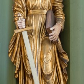 Zdjęcie nr 1: Rzeźba drążona ustawiona na niewielkim, prostopadłościennym postumencie. Święty Paweł został przedstawiony całopostaciowo, w pozycji stojącej, w lekkim kontrapoście.  W prawej ręce opuszczonej wzdłuż tułowia trzyma zamkniętą księgę, prawą wspiera na mieczu. Święty został przedstawiony jako dojrzały mężczyzna o wyrazistych rysach twarzy – głęboko osadzonych oczach, krótkim i wąskim nosie oraz wystających kościach policzkowych. Na głowie dłuższe, kręcone włosy w kolorze ciemnobrązowym oraz kędzierzawa broda w tym samym kolorze. Wzrok postaci skierowany przed siebie, usta nieco rozwarte. Święty Paweł jest ubrany w złocone, bogato fałdowane szaty – sięgającą ziemi suknię z długimi, wąskimi rękawami przewiązaną w pasie oraz złoty płaszcz udrapowany wokół prawej ręki. Widoczne spod szat stopy postaci są bose. Figura polichromowana, partie szat złocone.
