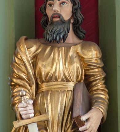 Zdjęcie nr 2: Rzeźba drążona ustawiona na niewielkim, prostopadłościennym postumencie. Święty Paweł został przedstawiony całopostaciowo, w pozycji stojącej, w lekkim kontrapoście.  W prawej ręce opuszczonej wzdłuż tułowia trzyma zamkniętą księgę, prawą wspiera na mieczu. Święty został przedstawiony jako dojrzały mężczyzna o wyrazistych rysach twarzy – głęboko osadzonych oczach, krótkim i wąskim nosie oraz wystających kościach policzkowych. Na głowie dłuższe, kręcone włosy w kolorze ciemnobrązowym oraz kędzierzawa broda w tym samym kolorze. Wzrok postaci skierowany przed siebie, usta nieco rozwarte. Święty Paweł jest ubrany w złocone, bogato fałdowane szaty – sięgającą ziemi suknię z długimi, wąskimi rękawami przewiązaną w pasie oraz złoty płaszcz udrapowany wokół prawej ręki. Widoczne spod szat stopy postaci są bose. Figura polichromowana, partie szat złocone.
