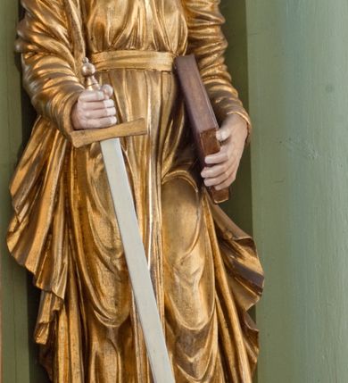 Zdjęcie nr 3: Rzeźba drążona ustawiona na niewielkim, prostopadłościennym postumencie. Święty Paweł został przedstawiony całopostaciowo, w pozycji stojącej, w lekkim kontrapoście.  W prawej ręce opuszczonej wzdłuż tułowia trzyma zamkniętą księgę, prawą wspiera na mieczu. Święty został przedstawiony jako dojrzały mężczyzna o wyrazistych rysach twarzy – głęboko osadzonych oczach, krótkim i wąskim nosie oraz wystających kościach policzkowych. Na głowie dłuższe, kręcone włosy w kolorze ciemnobrązowym oraz kędzierzawa broda w tym samym kolorze. Wzrok postaci skierowany przed siebie, usta nieco rozwarte. Święty Paweł jest ubrany w złocone, bogato fałdowane szaty – sięgającą ziemi suknię z długimi, wąskimi rękawami przewiązaną w pasie oraz złoty płaszcz udrapowany wokół prawej ręki. Widoczne spod szat stopy postaci są bose. Figura polichromowana, partie szat złocone.
