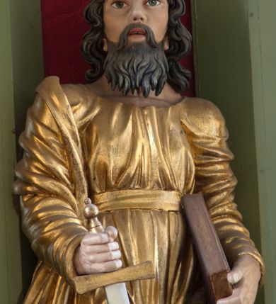 Zdjęcie nr 4: Rzeźba drążona ustawiona na niewielkim, prostopadłościennym postumencie. Święty Paweł został przedstawiony całopostaciowo, w pozycji stojącej, w lekkim kontrapoście.  W prawej ręce opuszczonej wzdłuż tułowia trzyma zamkniętą księgę, prawą wspiera na mieczu. Święty został przedstawiony jako dojrzały mężczyzna o wyrazistych rysach twarzy – głęboko osadzonych oczach, krótkim i wąskim nosie oraz wystających kościach policzkowych. Na głowie dłuższe, kręcone włosy w kolorze ciemnobrązowym oraz kędzierzawa broda w tym samym kolorze. Wzrok postaci skierowany przed siebie, usta nieco rozwarte. Święty Paweł jest ubrany w złocone, bogato fałdowane szaty – sięgającą ziemi suknię z długimi, wąskimi rękawami przewiązaną w pasie oraz złoty płaszcz udrapowany wokół prawej ręki. Widoczne spod szat stopy postaci są bose. Figura polichromowana, partie szat złocone.
