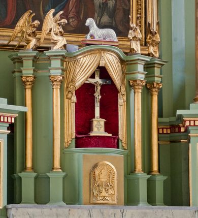 Zdjęcie nr 1: Tabernakulum z tronem wystawienia, jednokondycyjne o formach architektonicznych i wklęsło-wypukłym licu. Ujęte ustawionymi ukośnie, zdwojonymi, złoconymi kolumienkami wspartymi na wysokich cokołach. Kapitele kolumn kompozytowe, dźwigają odcinki pełnego belkowania wyłamanego w osiach podpór. Powyżej pełnoplastyczne, złocone figury klęczących aniołów. Drzwiczki tabernakulum złocone w kształcie stojącego prostokąta zamkniętego łukiem pełnym. Na licu płaskorzeźbiony kielich z hostią na tle glorii promienistej oraz kłosów zboża i winnych gron. Tron wystawienia o linii wypukłej z niszą ujętą płaskorzeźbioną, złoconą kotarą dekorowaną chwostami oraz muszlami palmetowymi. Tło wyłożone aksamitną, czerwoną tkaniną. W zwieńczeniu srebrzony Baranek Eucharystyczny spoczywający na księdze. Struktura tabernakulum malowana w odcieniu seledynowej zieleni, ornamentyka i załamania profili architektonicznych złocone.

